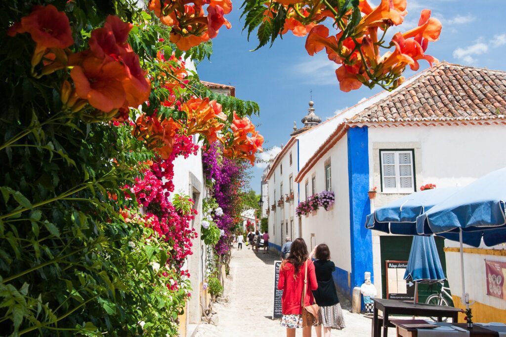 Colorful Mediterranean village street with flowers, tourists, and outdoor cafés, creating a picturesque scene.