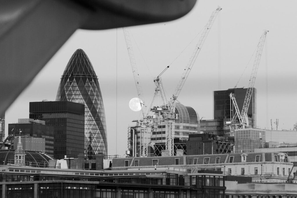 London Gherkin skyscraper, cranes, and moon in a modern cityscape at night.