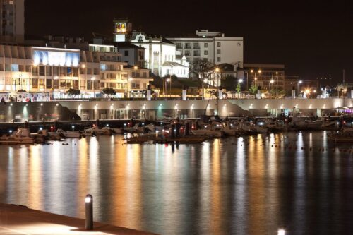 Tranquil coastal cityscape at night with shimmering lights reflecting on calm water.