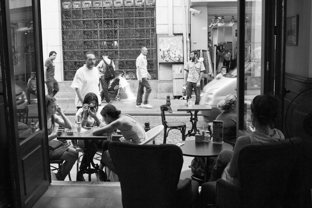 Lively urban café scene with patrons inside and bustling pedestrians on the street outside.