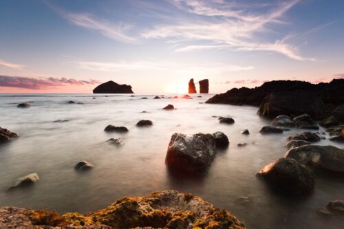 Tranquil coastal sunrise or sunset with rock formations and calm ocean view.