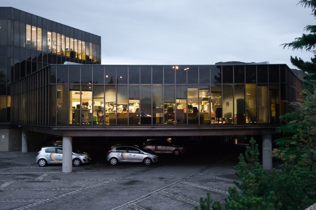 Sleek glass building with cantilevered design, parking below, warm office space, and green surroundings.