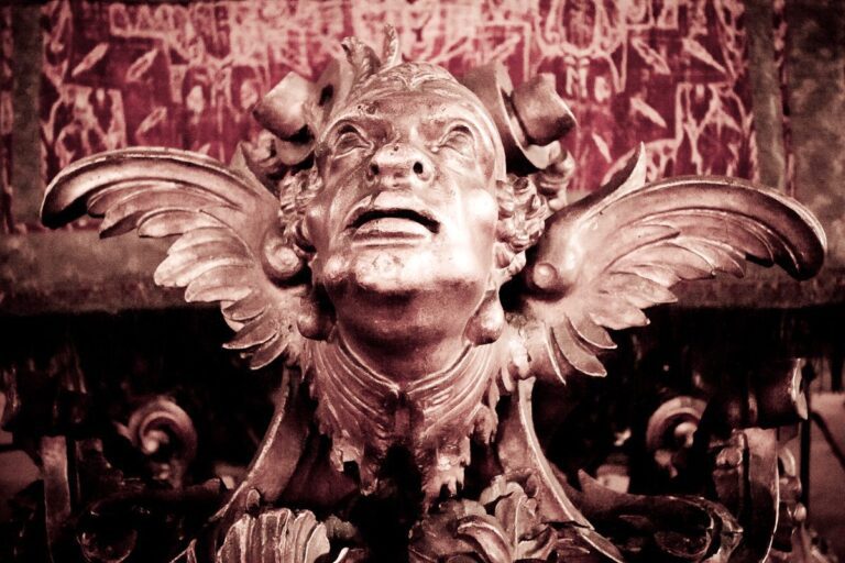 Baroque sculpture with dramatic face, wings, and ornate details against rich red background.