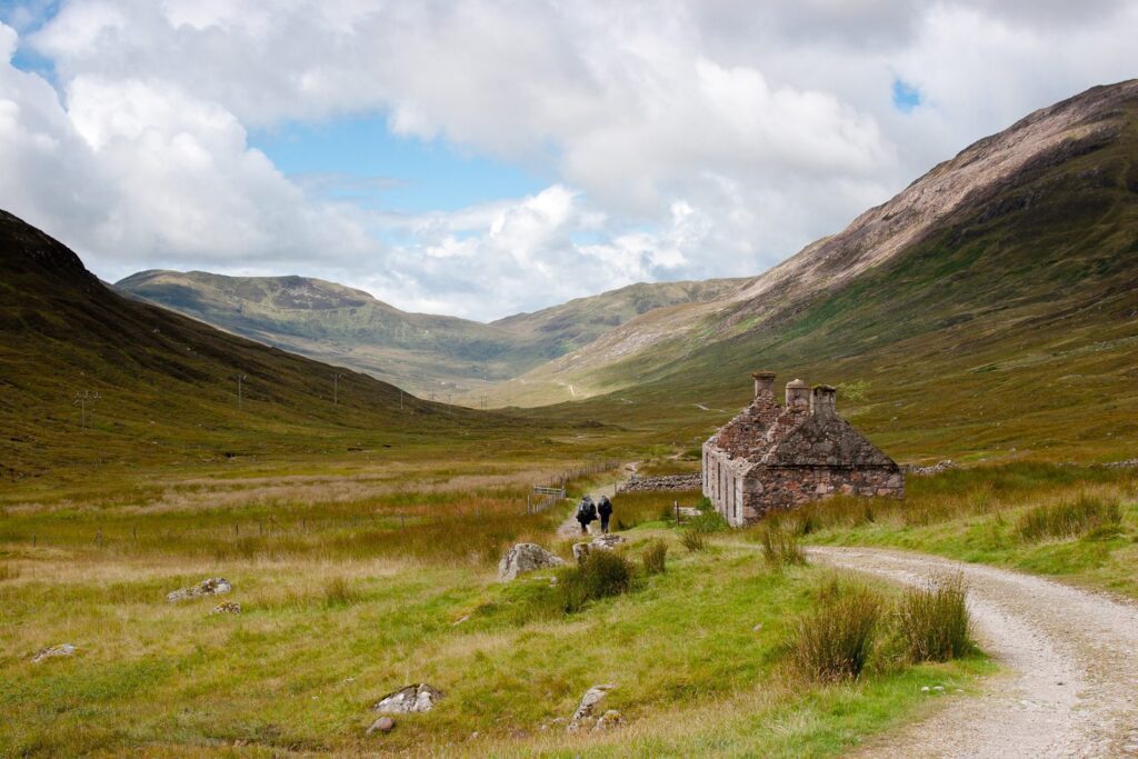 Tranquil mountain valley with stone cottage, lush fields, rolling hills, and blue sky.