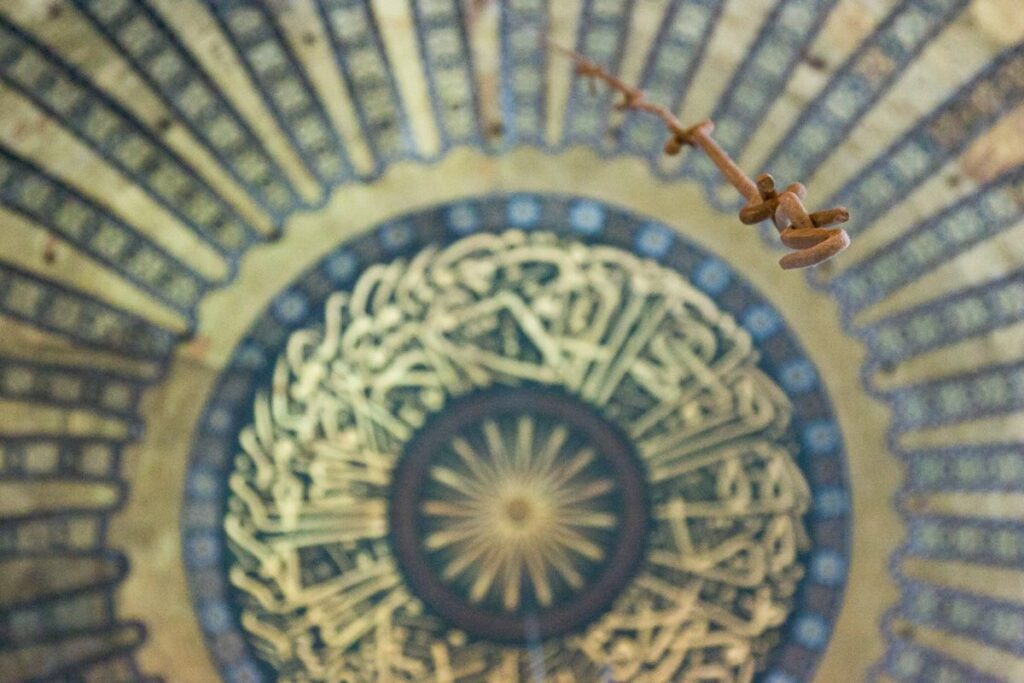 Intricately designed mosque dome with golden sunburst, Arabic calligraphy, and a bronze chandelier.