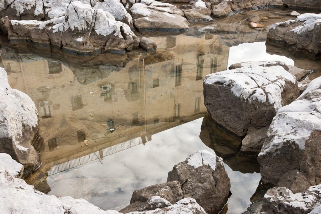 Tranquil reflection of old building in calm water with snow-covered rocks.