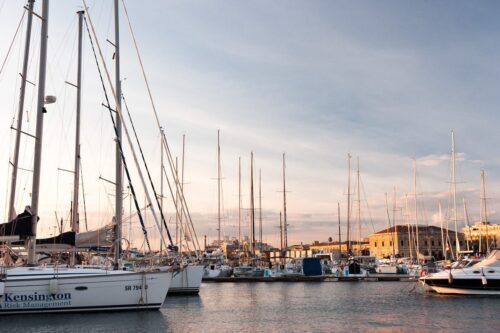 Tranquil marina at sunset with sailboats, yachts, and traditional buildings reflecting in calm waters.