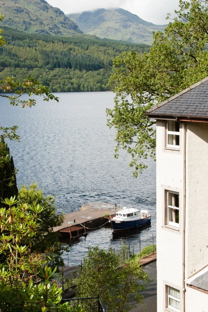 Tranquil lakeside retreat with white building, dock, boat, lush foliage, forested hills, and misty mountains.